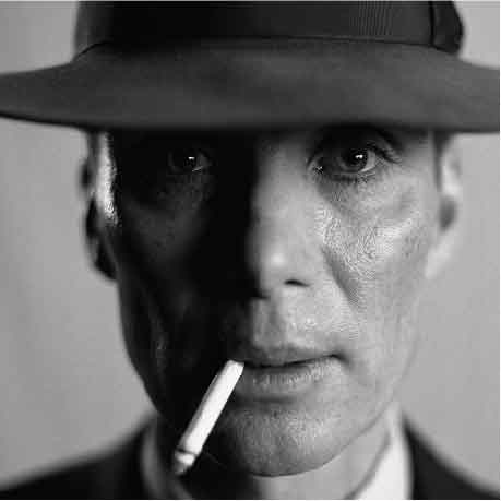 A close-up of Cillian Murphy portraying Robert Oppenheimer, the close up is in black and white. Oppenheimer is staring with no expression he has a cigarette dangling from his mouth.